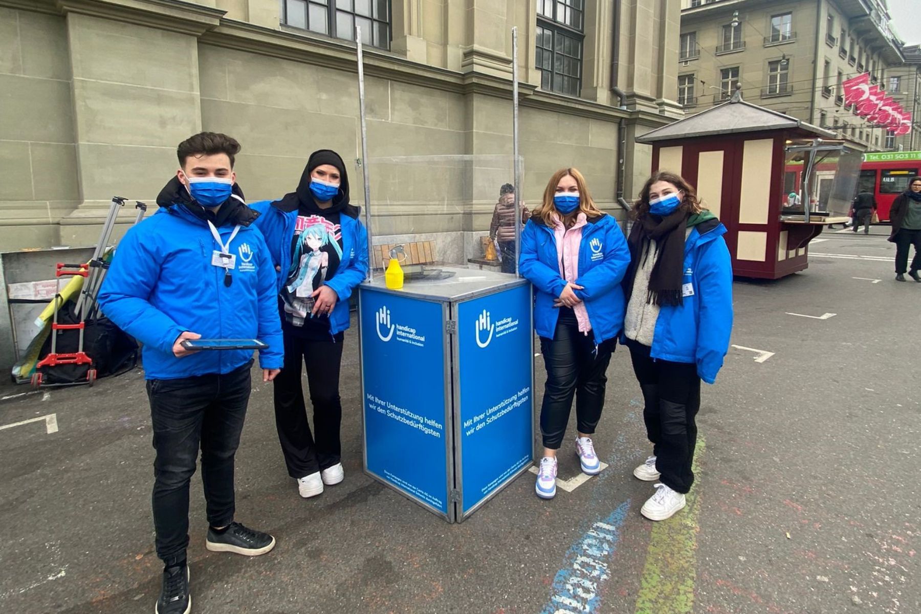 Des jeunes dialogueurs habillé avec des vestes bleues aux couleurs de HI sont devant la gare de Berne avec un stand HI.