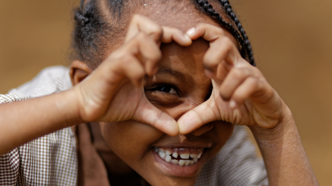 Une petite fille qui souris et qui fait un coeur avec ses mains.