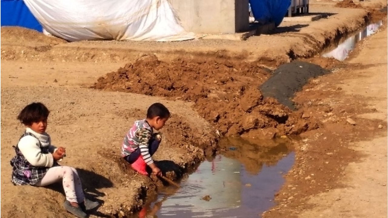 Zwei kleine Kinder spielen am Rande einer Wasserrinne im Zeltlager Khazer