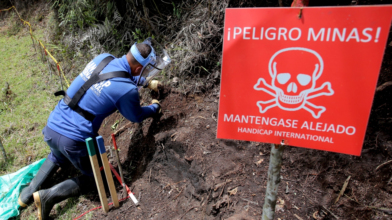 Ein rotes Schild warnt vor den Minen. Danaben kniet ein Entminer von Handicap International und sucht gezielt nach Minen.
