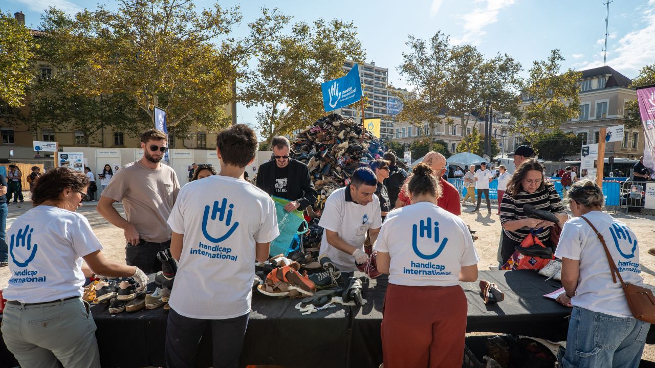 Personnes portant un t-shirt Handicap International derriere un stand sur une place à Lyon. Derrière on voit une pyramide de chaussures.