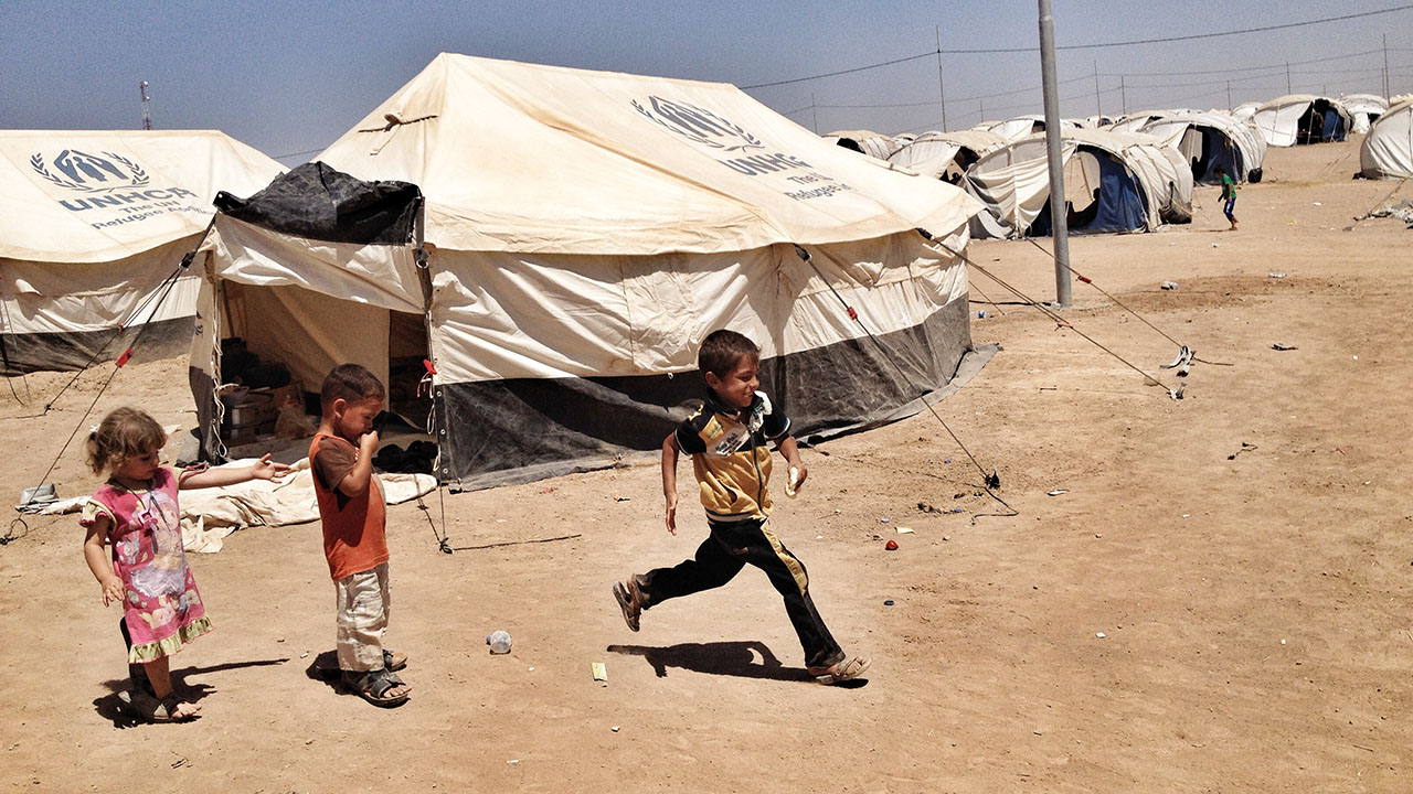3 Kinder, zwei Jungs und ein Mädchen spielen zusammen im Flüchtlingslager Khazir Camp. Im Hintergrund sind weiße Stoffhütten zu sehen.