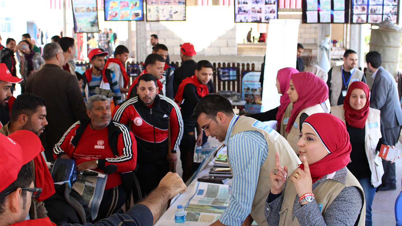 Viele Menschen stehen um einen Informationsstand in Gaza. Es sind Flyer und andere Dokumente ausgelegt. Zwei Expert*innen von HI sind mit den Besuchenden im Gespräch.