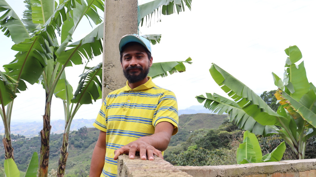 Portrait d'un homme qui s'appuie sur un muret et sourit à la caméra. Derrière lui, on voit des branches de bananiers et on devine au loin des collines verdoyantes.