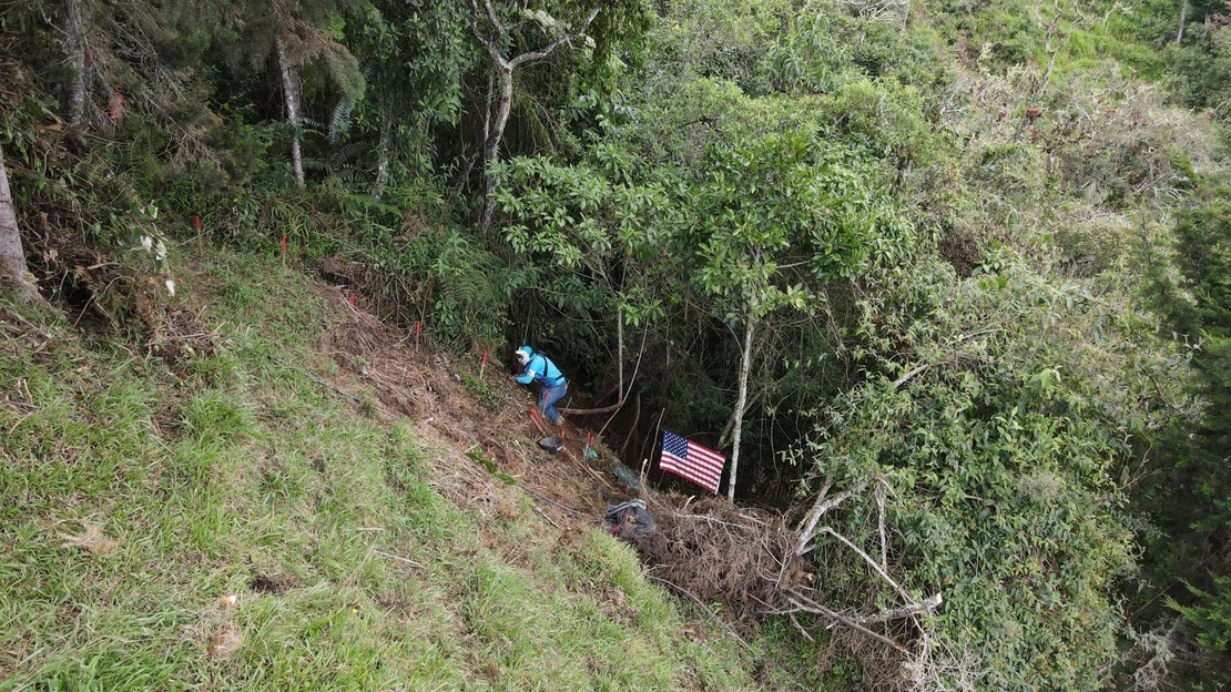 Un démineur est accroupi sur une pente raide et démine le terrain devant lui. Autour de lui, une forêt luxuriante. Un drapeau des États-Unis figure à côté de lui.