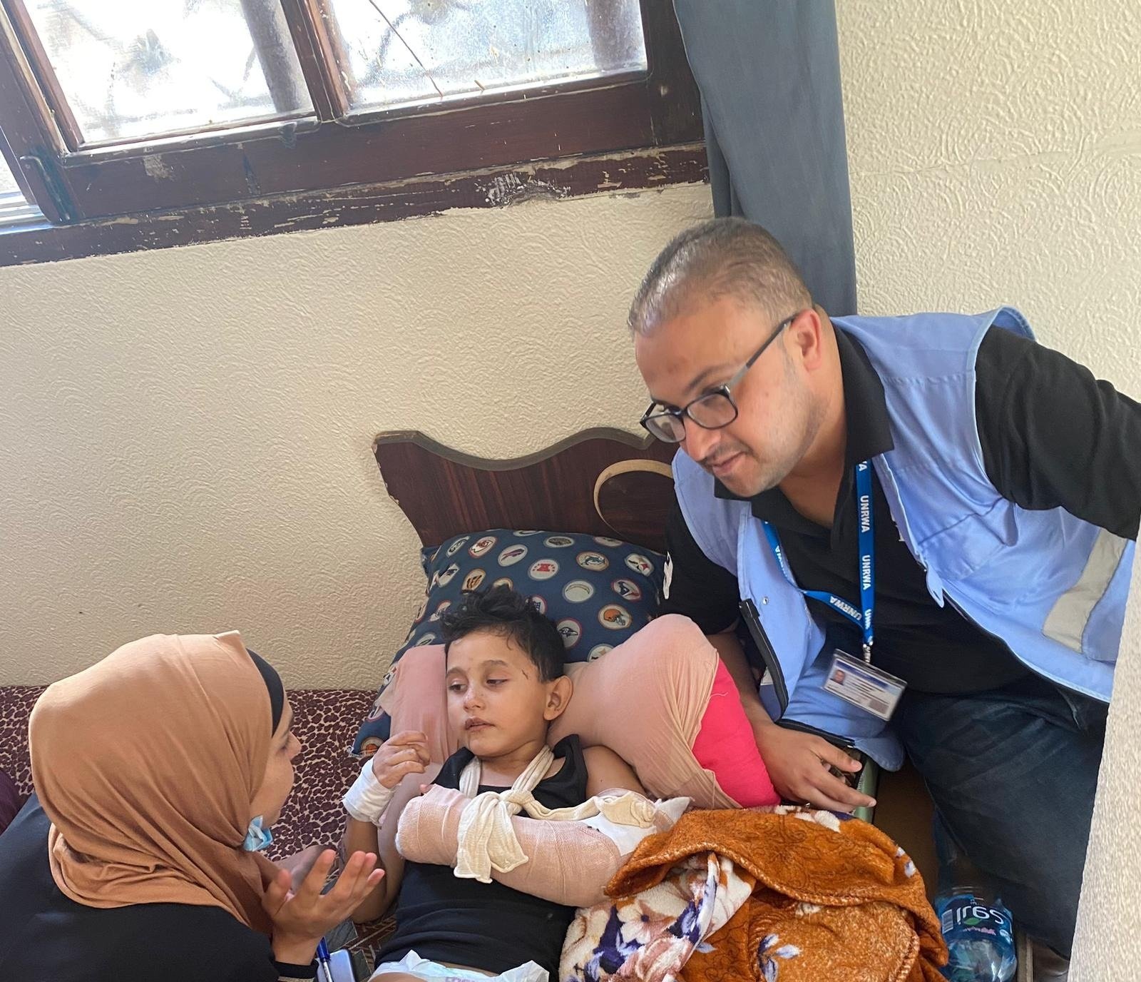 Un enfant est blessé à Gaza. Il est couché dans un lit avec une femme et un homme autour de lui. Son bras est dans un platre. 