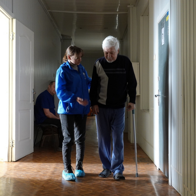 Ein älterer Begünstigter in der Ukraine steht in einem Flur mit einer Krücke. Eine weibliche HI-Angestellte hält seinen Arm fest. 